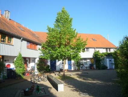 Ein graues Haus mit rotem Dach. In der Mitte ein Baum. 